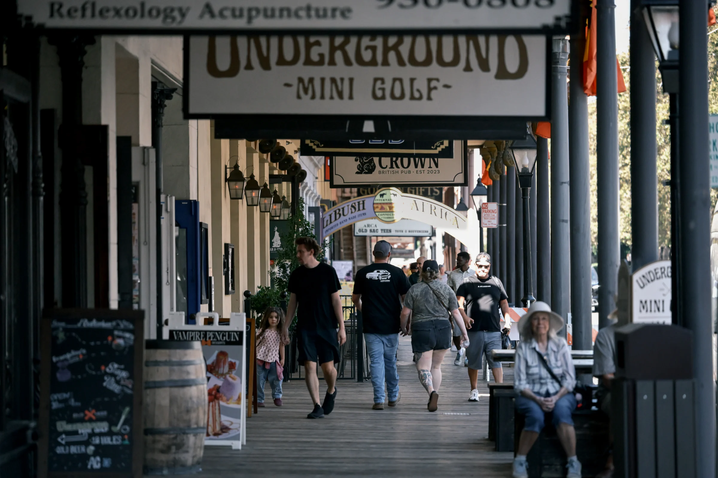 People walking and sitting beneath hanging shop signs along a column-lined walkway