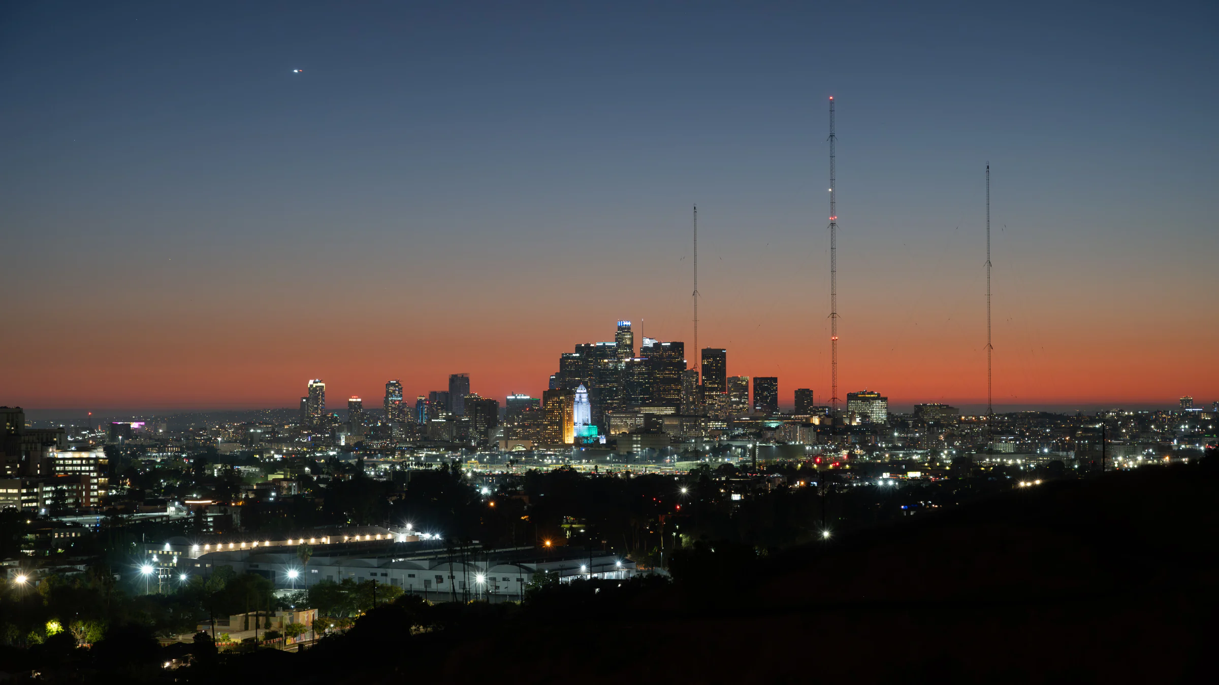 Illuminated city skyline and radio towers beneath an orange and blue dusk sky