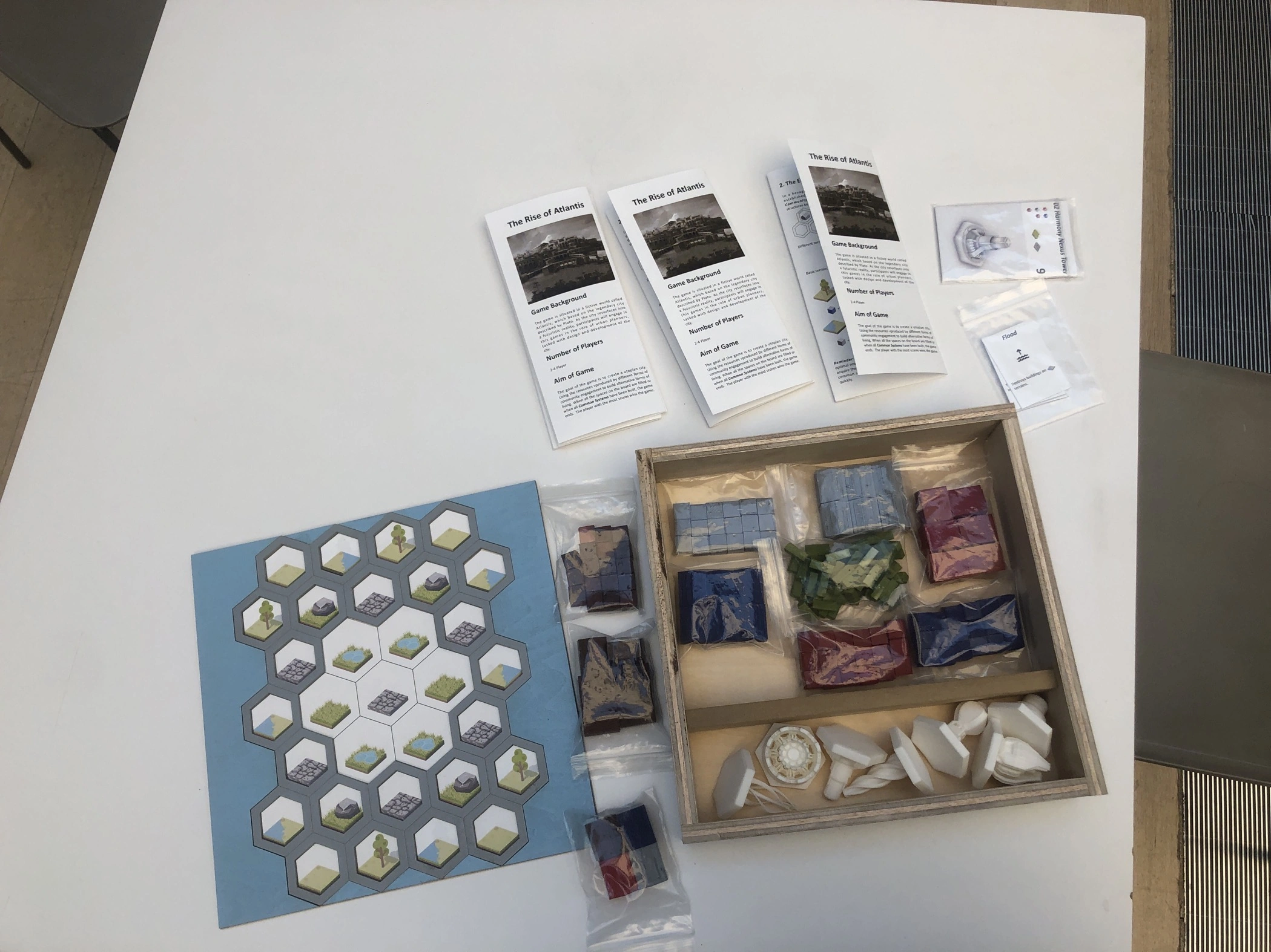Archival tabletop photograph of the Atlantis board, printed rules and cards, bagged colored components and seven white architectural pieces in a wooden box